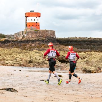 Two men running past Archirondel tower taking part in Breca Swimrun