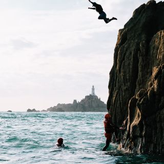 A man leaping into the water from the the top of larger rock with Corbiere Lighthouse in background.