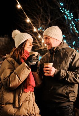 Couple smiling each other. They are wearing warm, wooly, coats are are surrounded by people and bright Christmas lights.